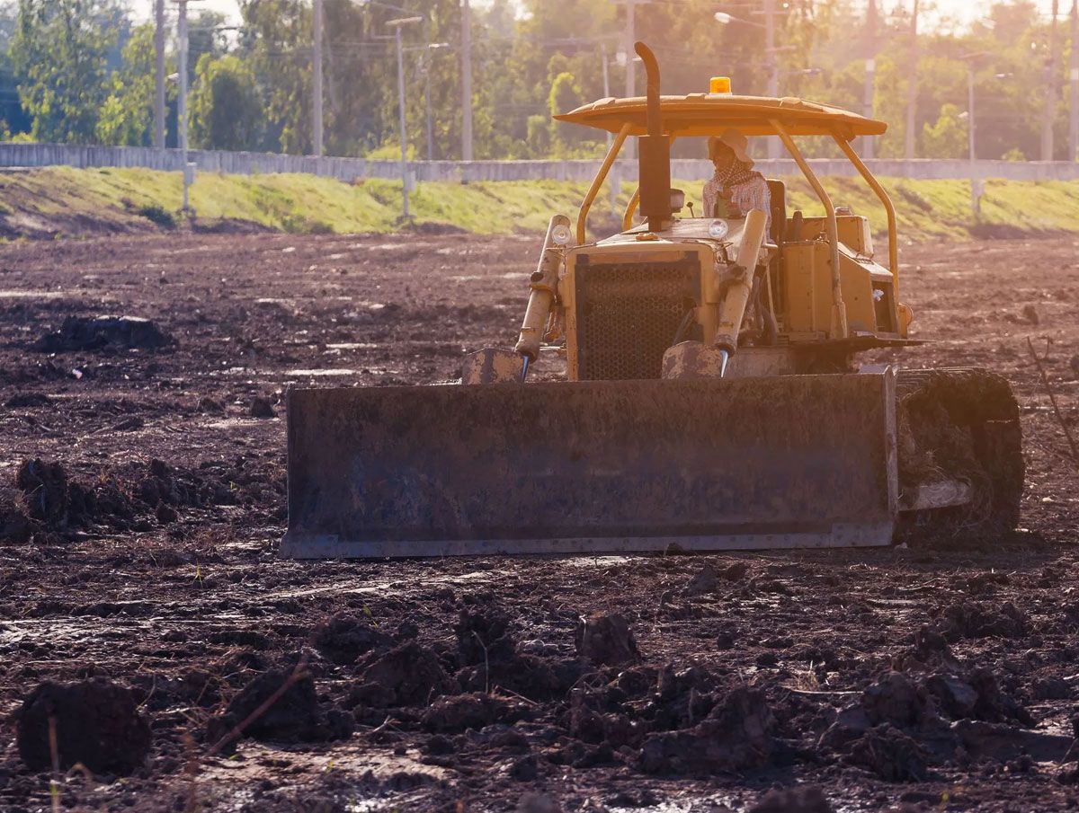 A yellow bulldozer sits on a dark, muddy field during a bright sunset, with trees visible in the background.