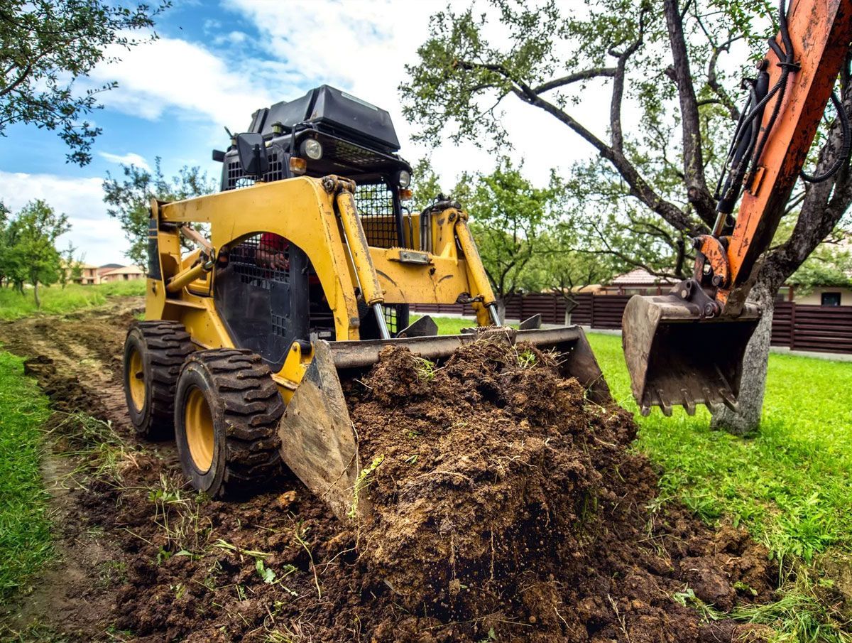 A yellow skid steer loader pushing a large pile of dirt, with a partial view of an excavator bucket in a grassy area.