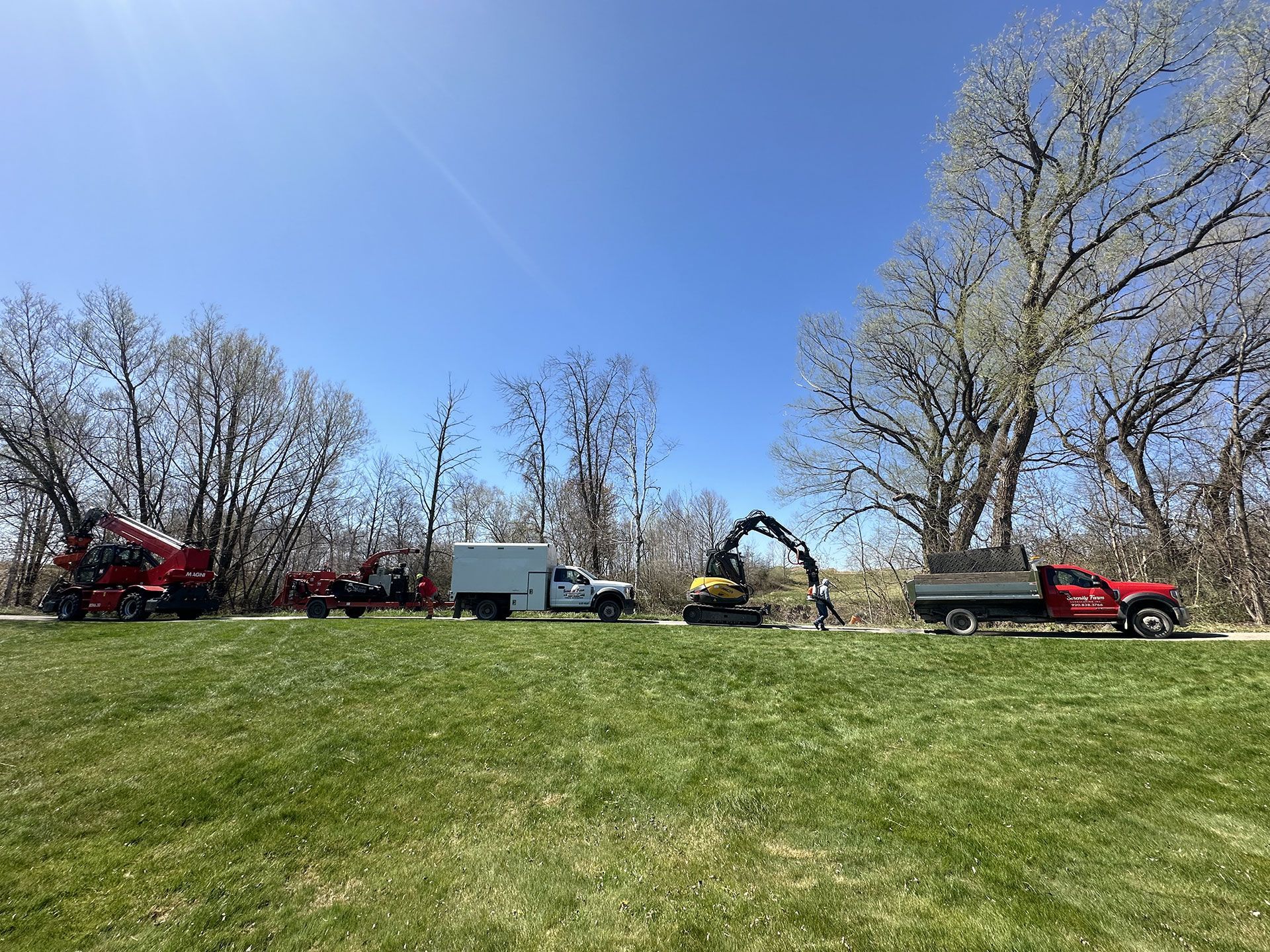 A work crew with heavy equipment, including a red crane, a service truck, and a yellow excavator, on a grassy field.