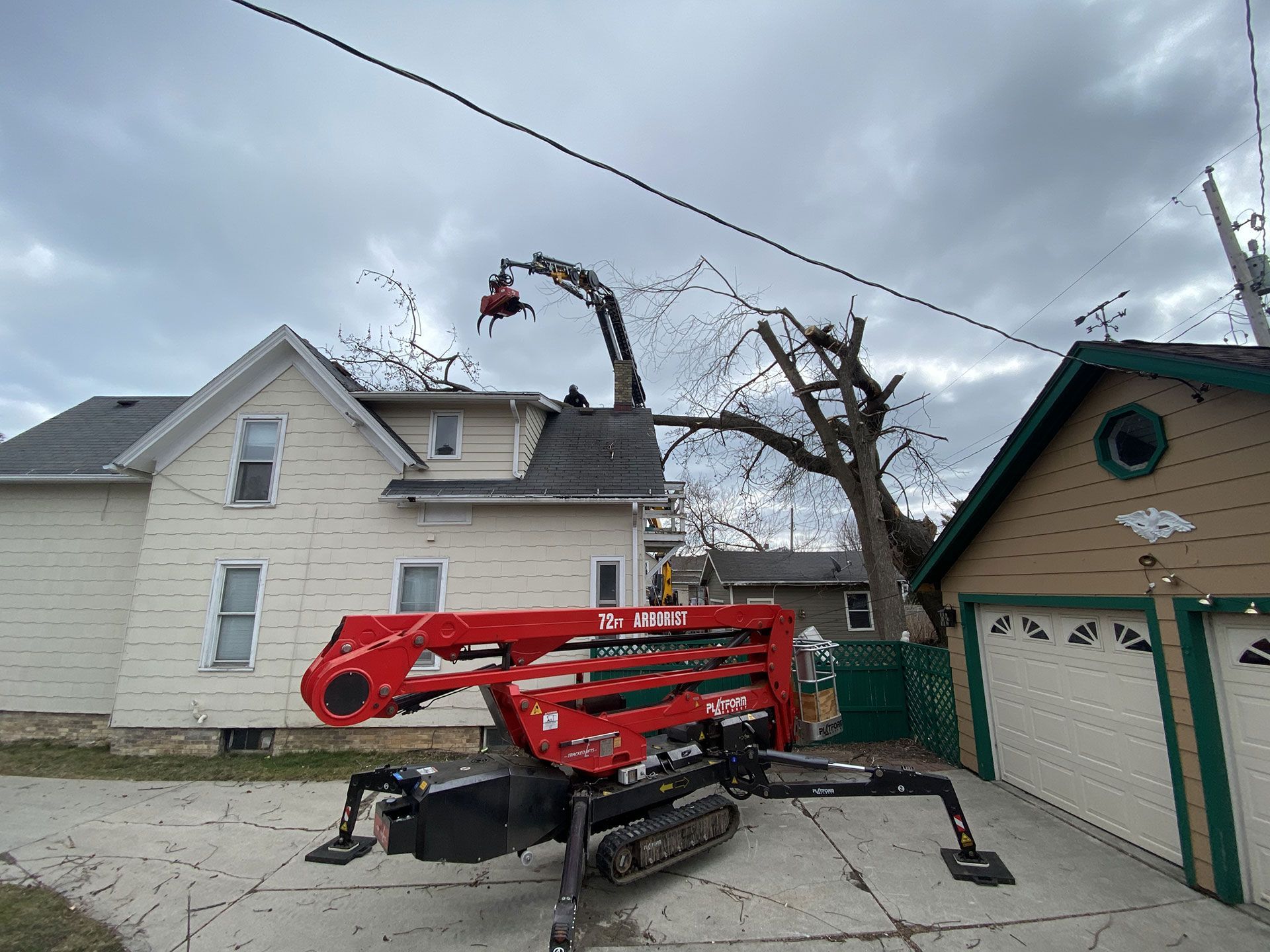 A red tracked lift operates in a driveway, extending an arm to remove tree limbs overhanging a two-story white house.