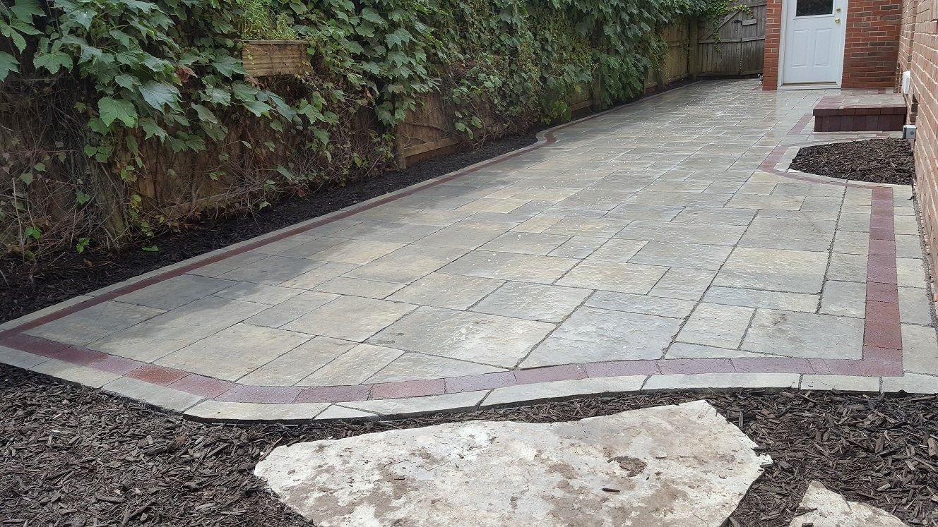 A paved patio made of light stone tiles with a dark red border, set in a yard with dark mulch and a stone stepping stone.