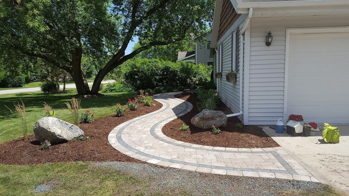A light-colored paver walkway curves through a mulched landscape bed toward the side of a white house with a garage.