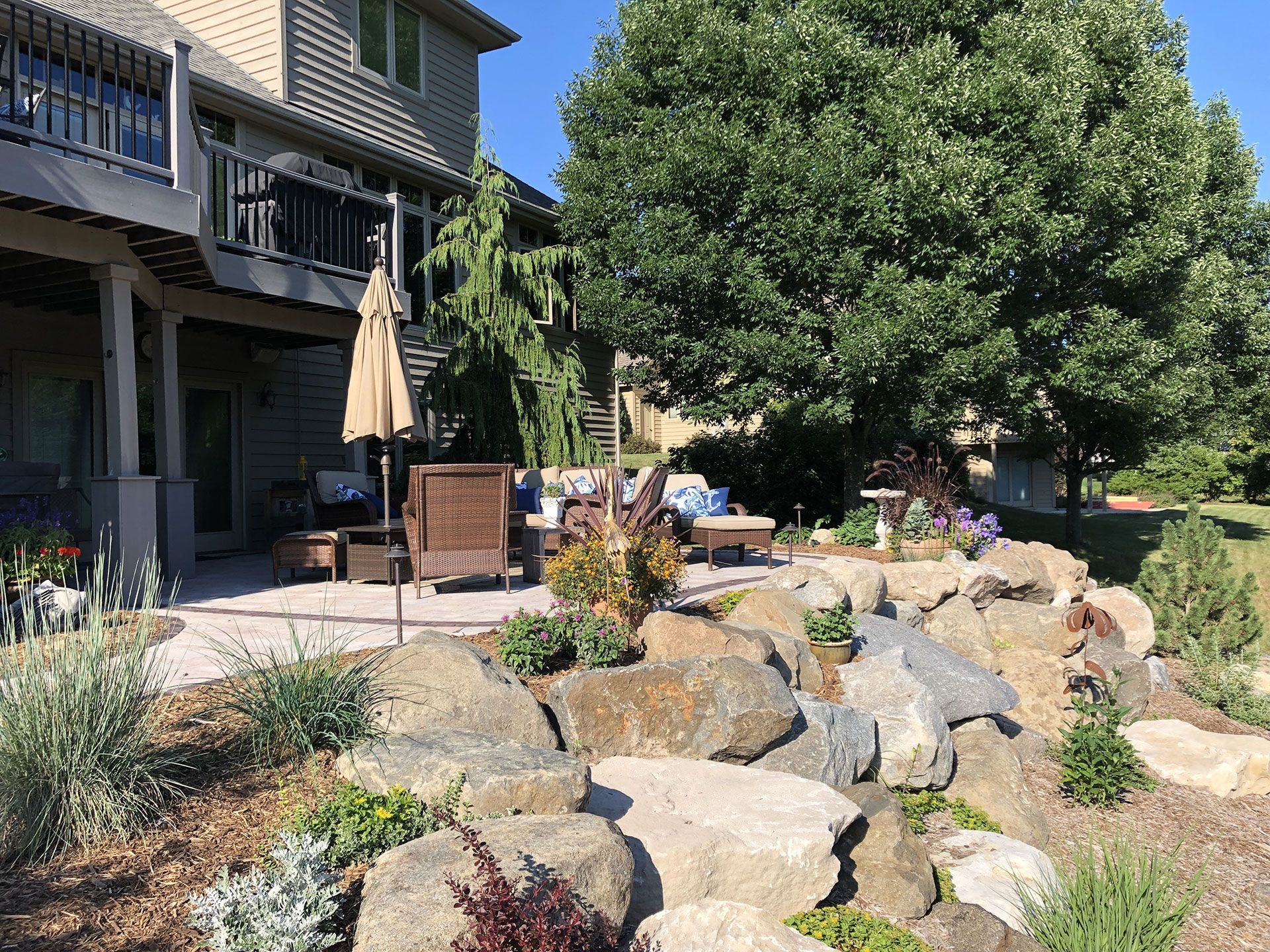 A patio with outdoor furniture under a tan umbrella, situated next to a stone retaining wall and a large leafy tree.