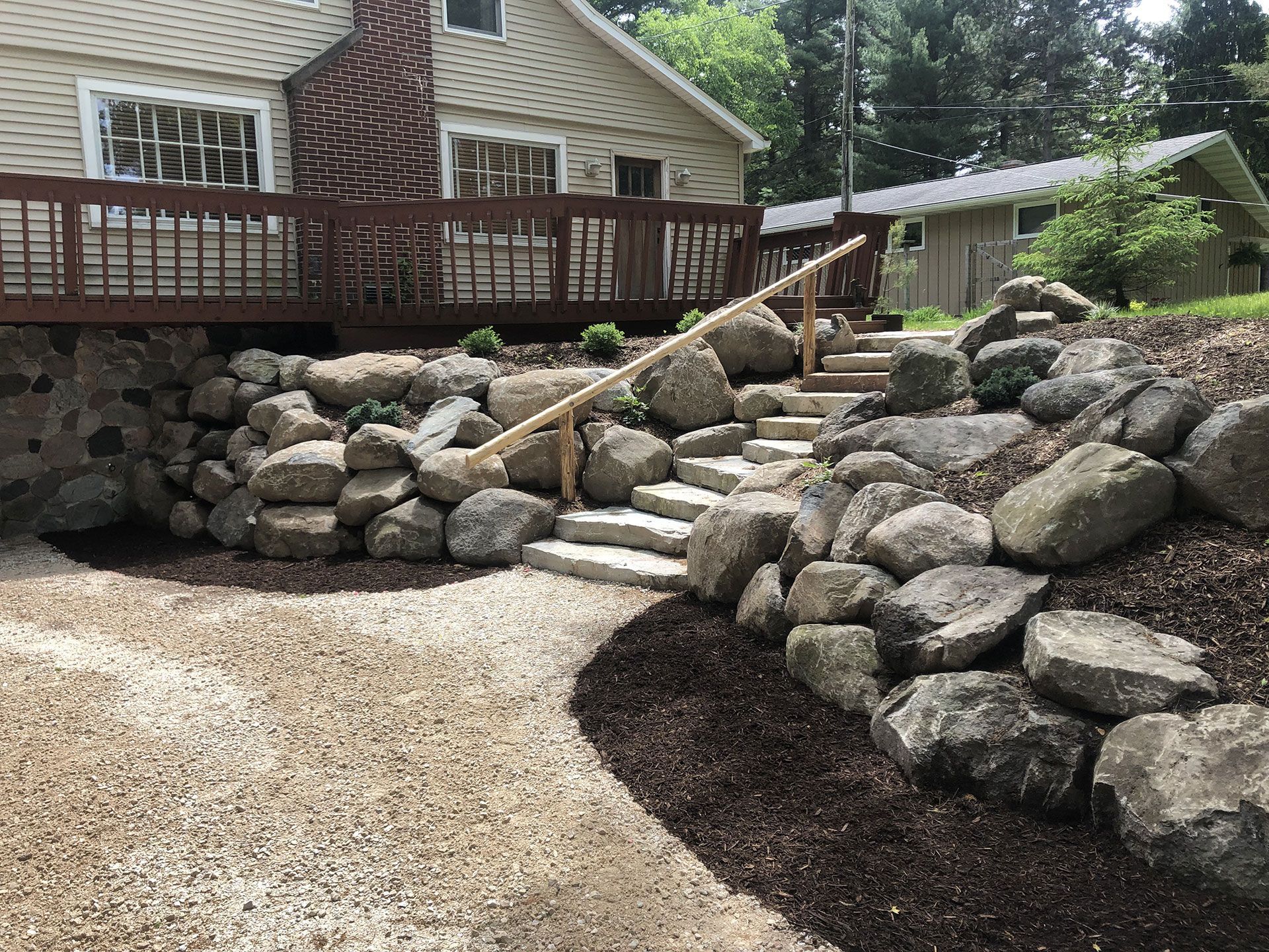 A stone staircase with a wooden handrail leads up a landscaped hill to a house deck, flanked by gravel and mulch beds.