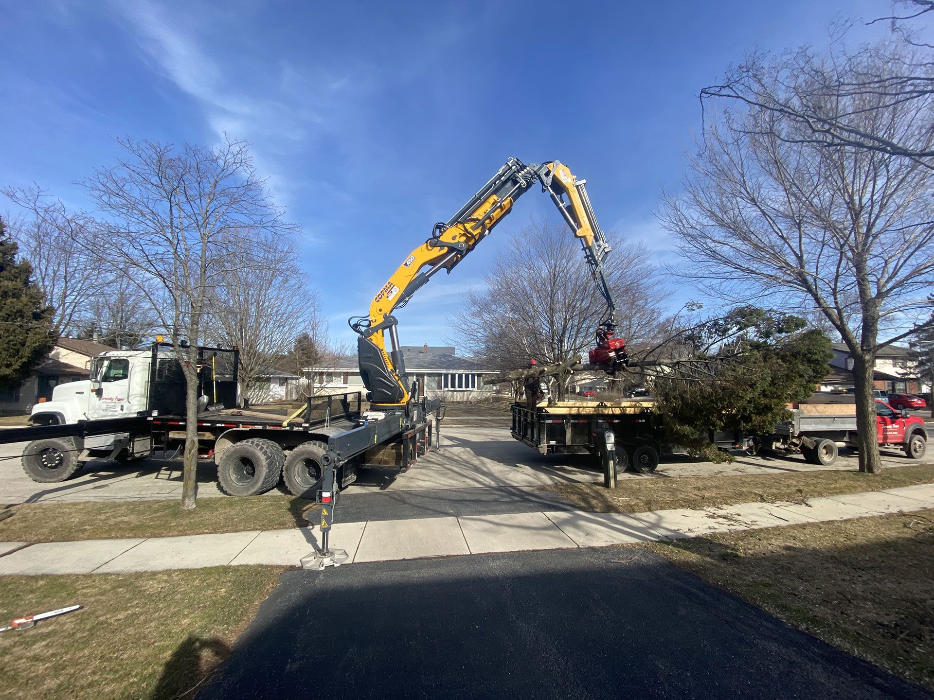 A yellow crane truck lifts branches into another truck on a sunny residential street.