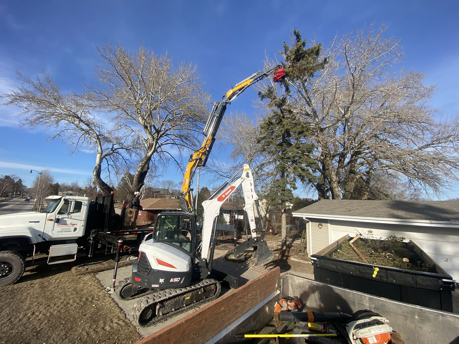A white Bobcat excavator with an extended boom attachment trims a tall evergreen tree near a truck and house.