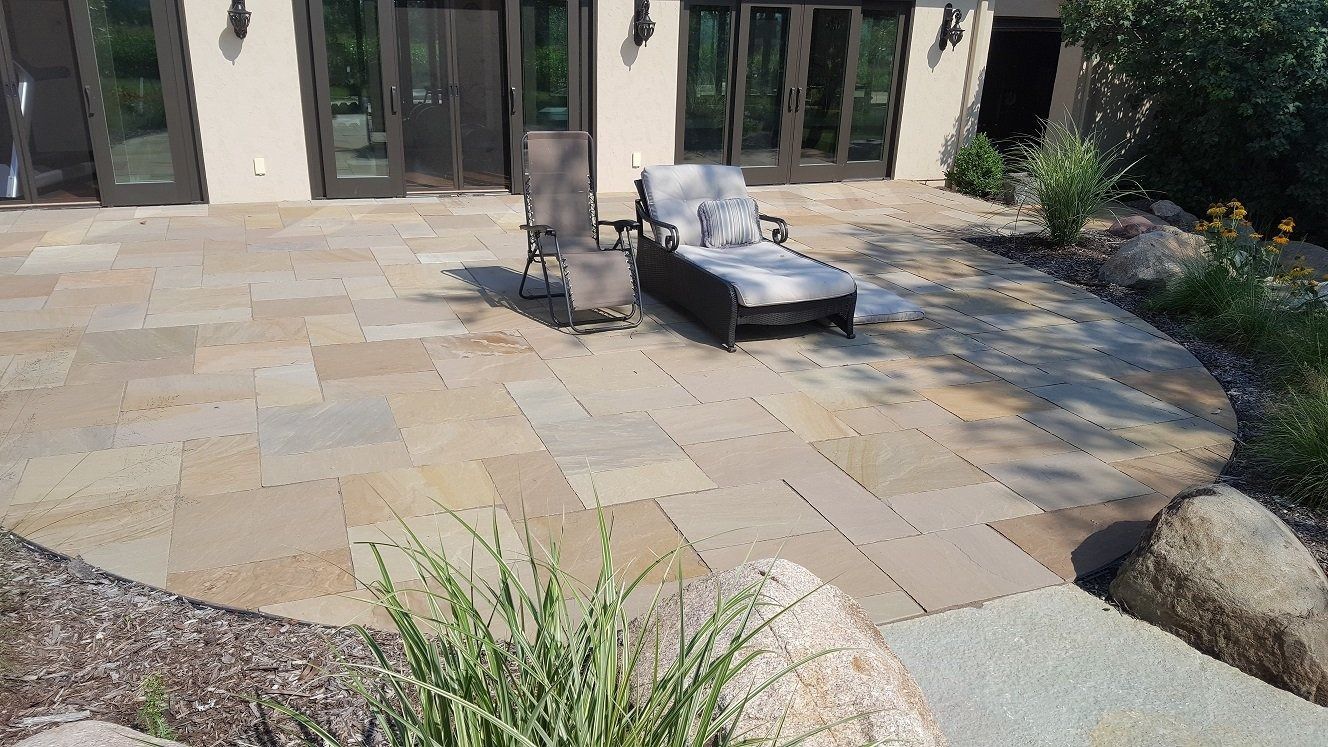 A stone patio with a lounge chair and recliner outside a house with glass doors, surrounded by landscaping and rocks.