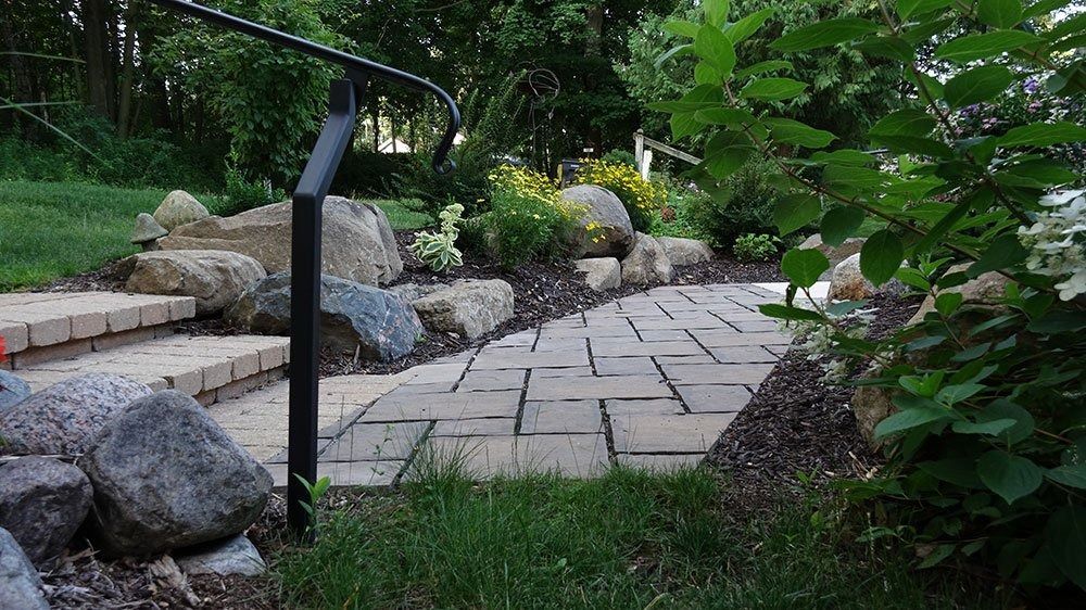 A paved stone walkway with stone steps and a black metal handrail leading through a garden with rocks and greenery.