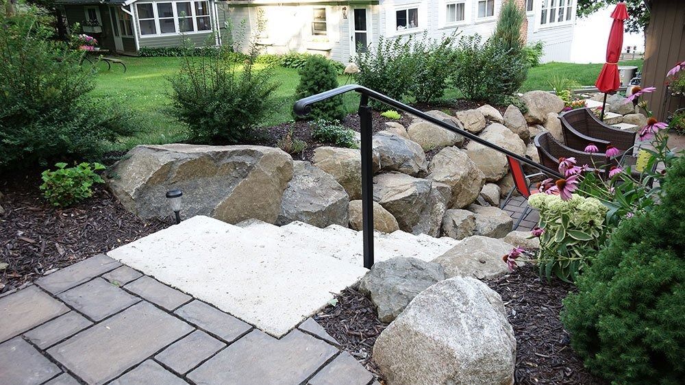 Stone steps with a black metal handrail leading up a landscape of large rocks and greenery to a house.