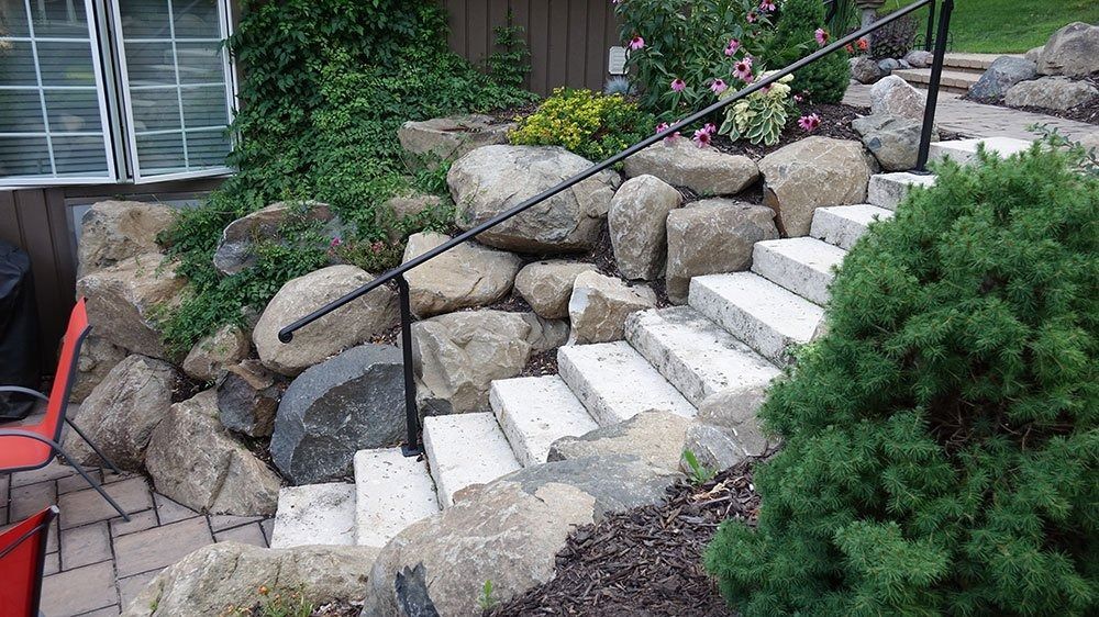 Stone stairs with a black metal handrail leading up a landscaped slope with large rocks and green shrubs.