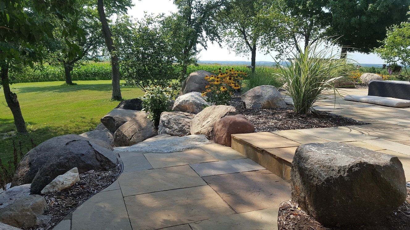 A stone patio walkway framed by large decorative boulders, garden plants, and trees under a sunny sky.