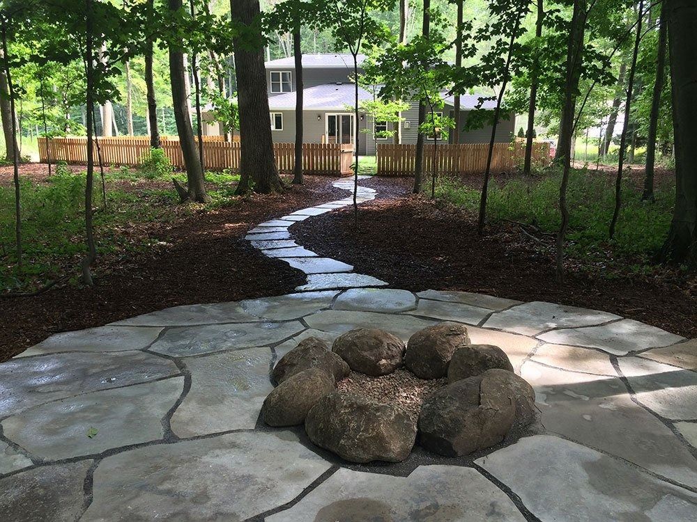 A stone fire pit on a circular patio leads to a winding flagstone path toward a house nestled in a wooded backyard.