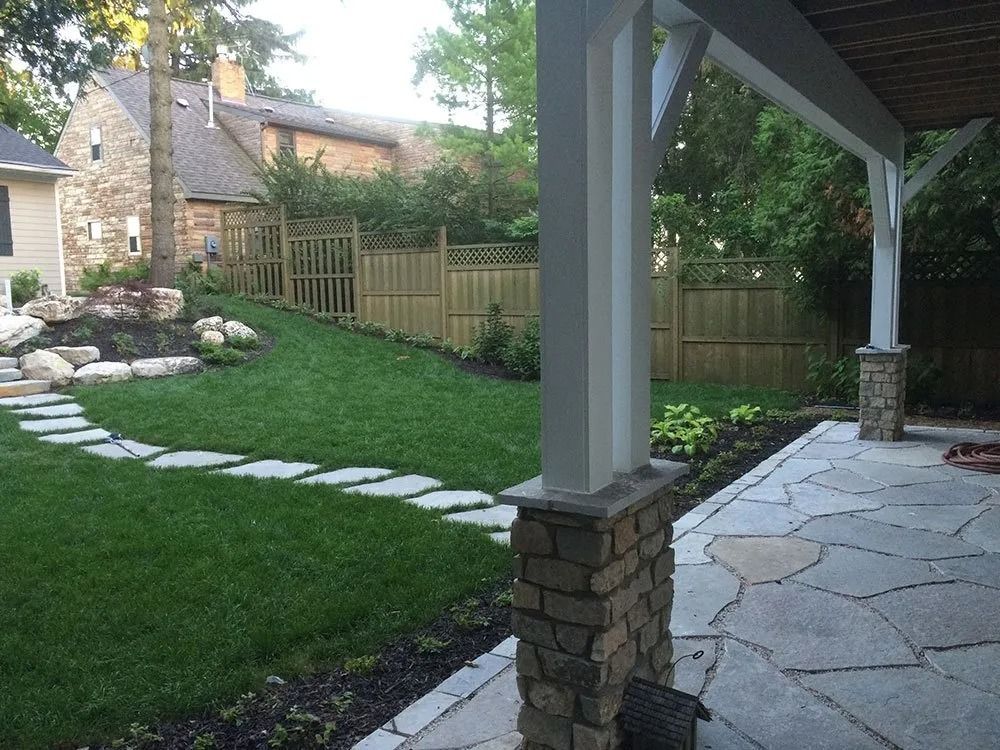 A stone patio under a roof, overlooking a grassy lawn with a stone stepping-stone path and a wooden fence.