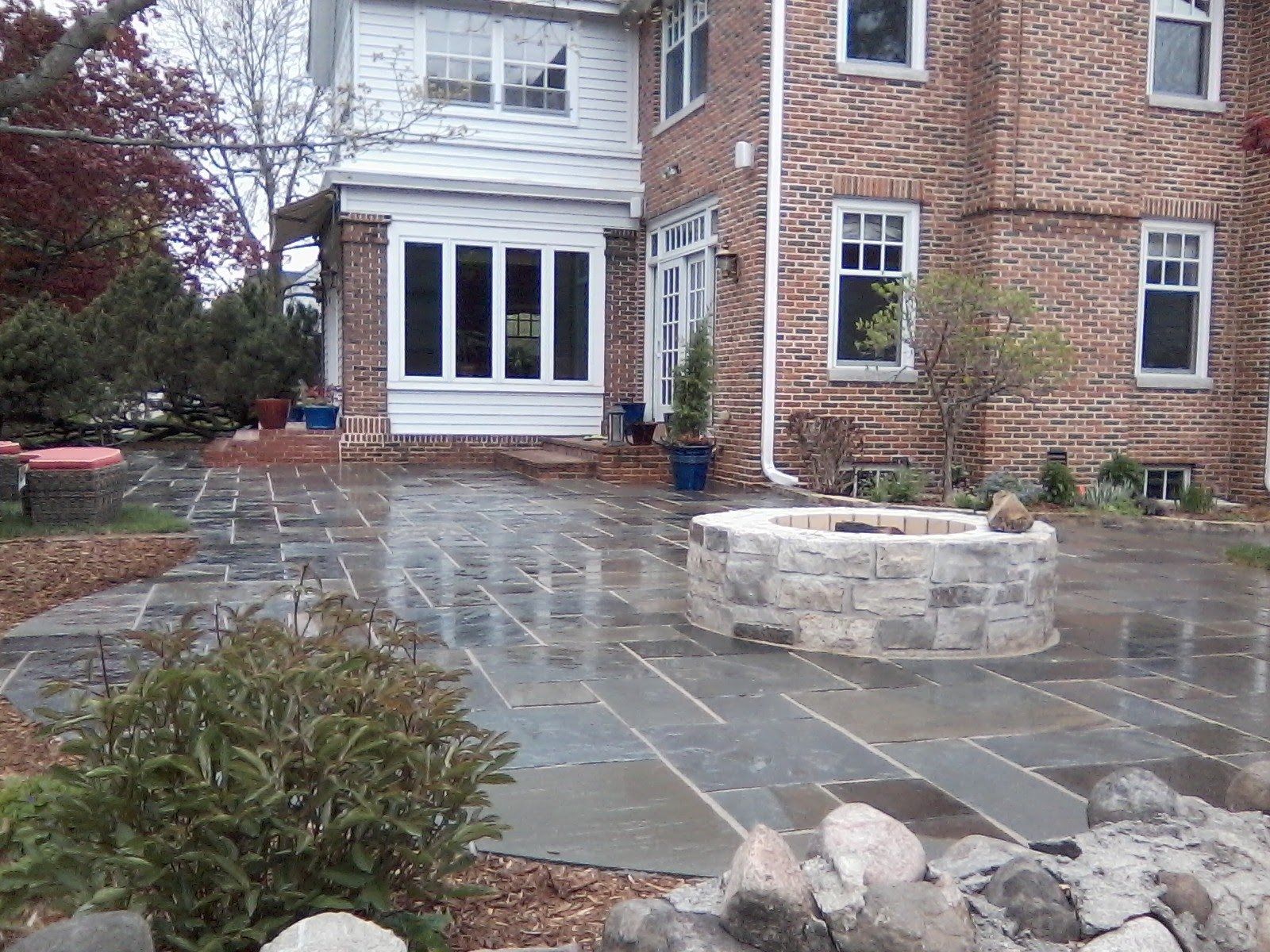 A stone patio with a round fire pit in front of a house with brick and white siding walls on a cloudy, wet day.