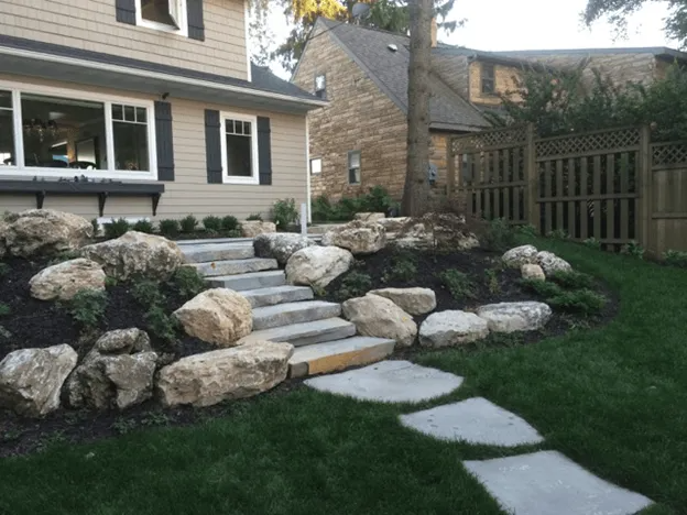 Stone steps lead up a landscaped hill to the entrance of a beige house with a stone-clad neighbor in the background.