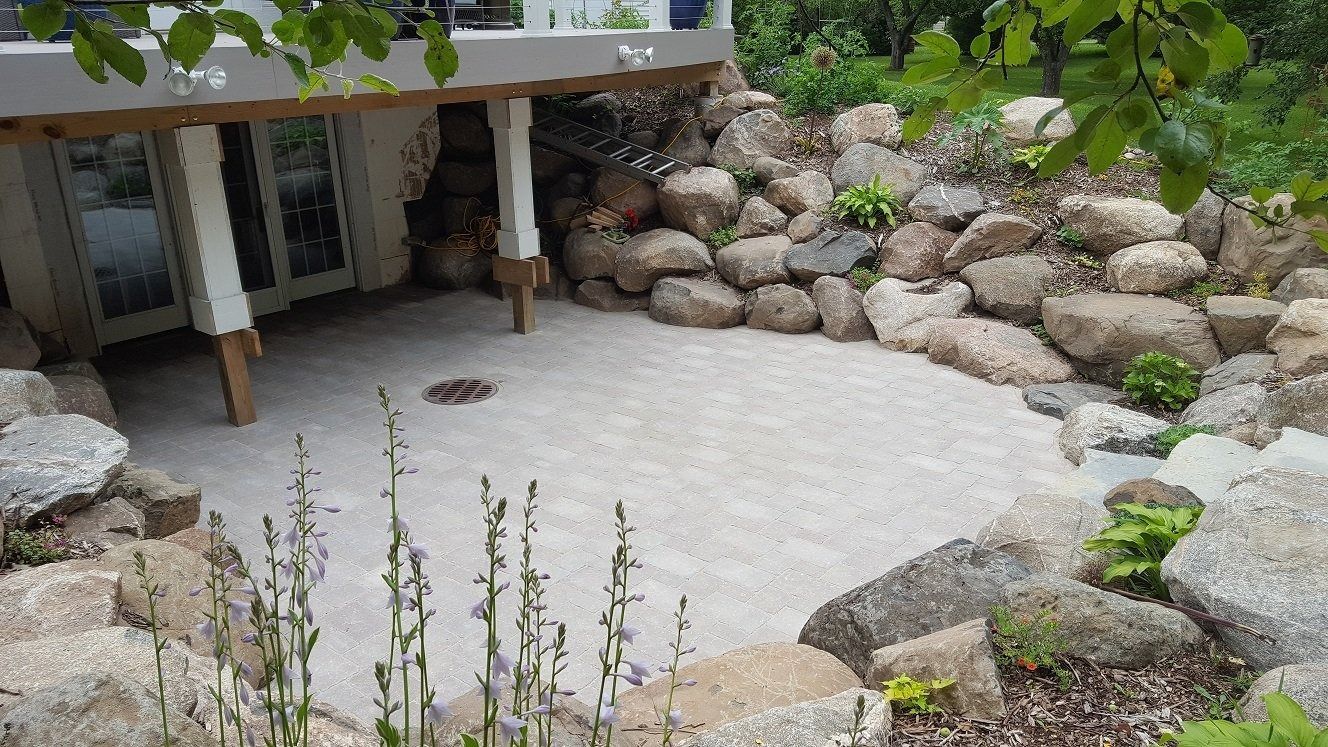 A gravel patio with a fire pit sits beneath a raised deck, surrounded by a natural rock retaining wall and lush greenery.