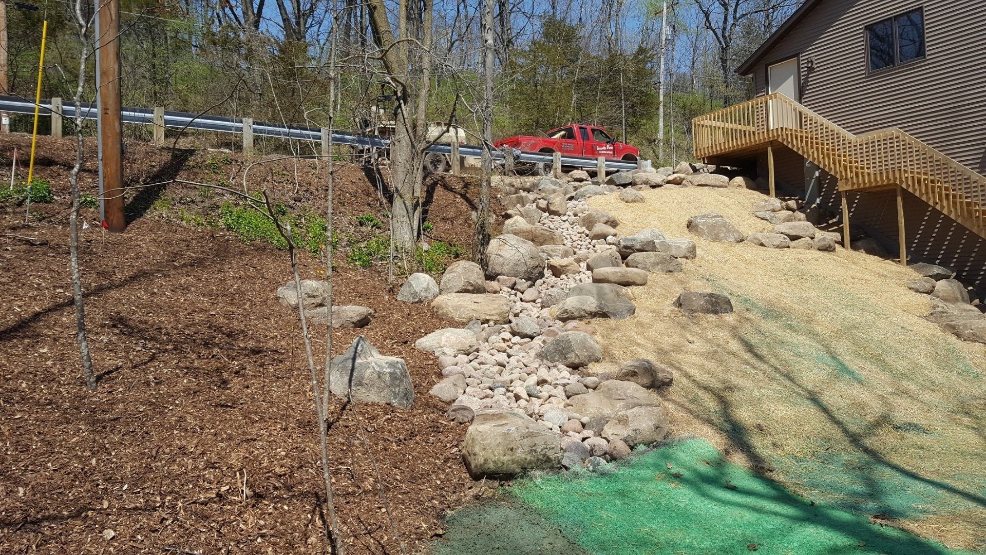 A sloped yard with a new rock drainage swale, mulch, and green erosion control netting leading to a wooden deck and house.