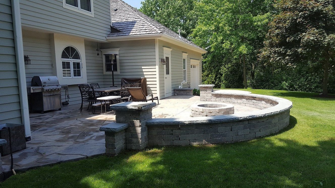 A backyard stone patio featuring a curved retaining wall with an integrated fire pit, adjacent to a gray house.