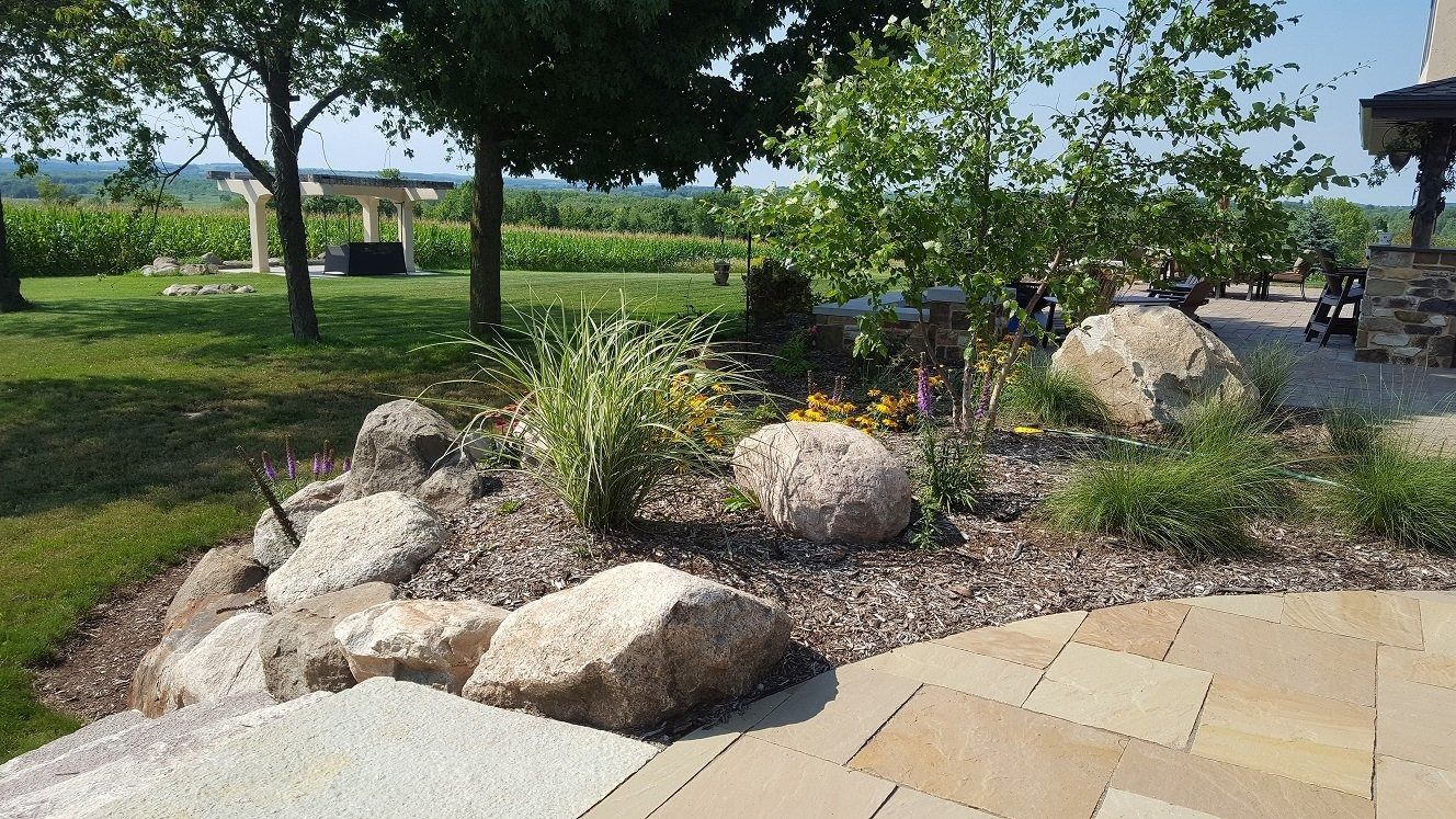 Landscaped garden bed with large boulders and shrubs, bordered by a stone patio and a lawn with a distant pergola.