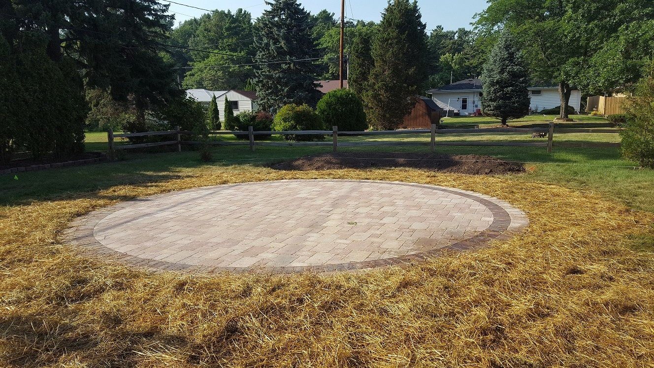 A circular area of leveled sand or gravel centered in a grass lawn, surrounded by dead leaves and trees in the background.