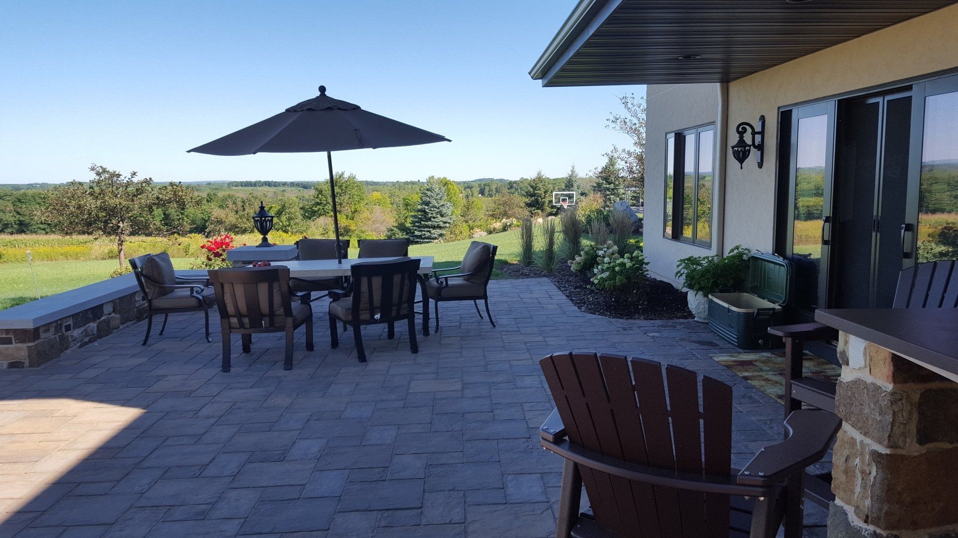 A stone patio with outdoor dining furniture and an umbrella, overlooking a scenic landscape under a clear blue sky.