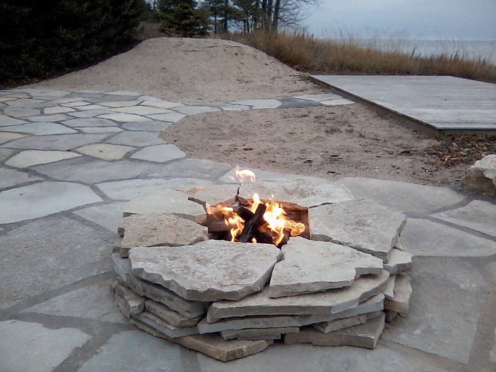 A stone fire pit with a burning fire, set on a natural stone patio near a sandy area and a body of water.