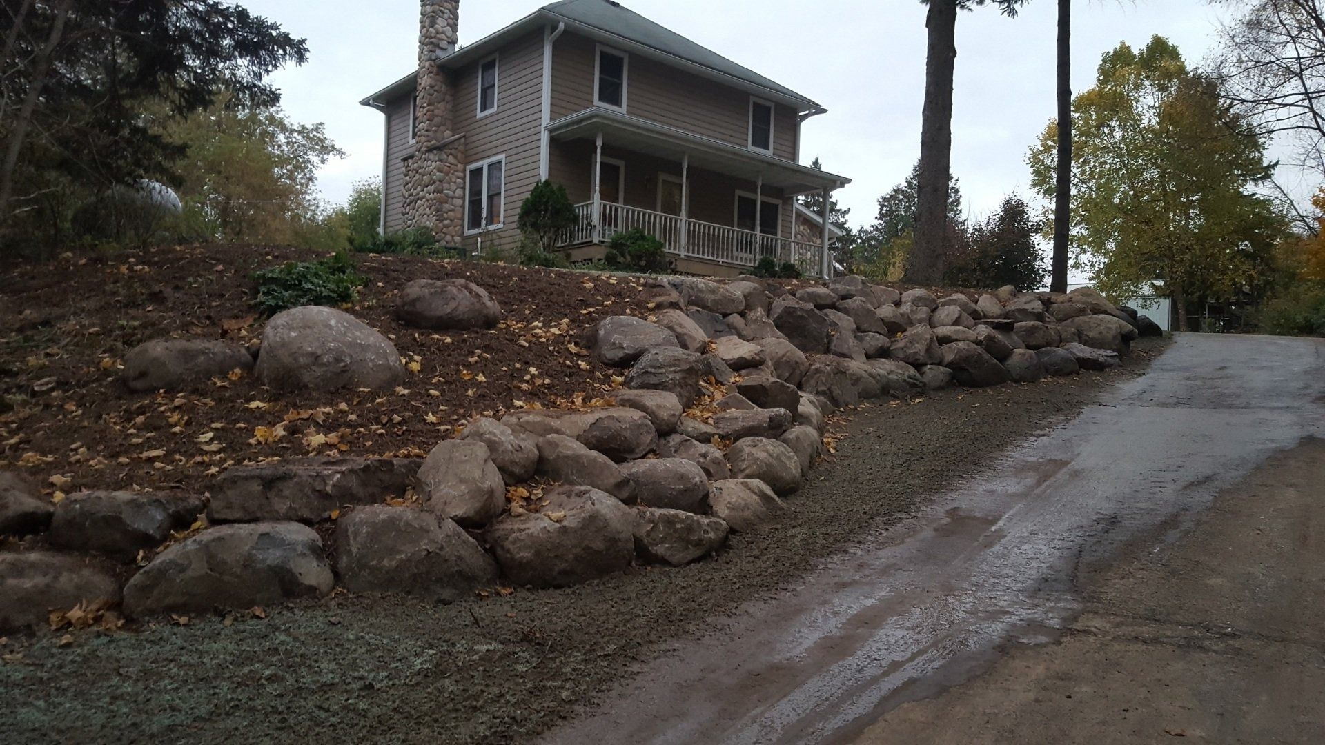 A large, multi-level boulder retaining wall sits in front of a tan, two-story house near a paved driveway.