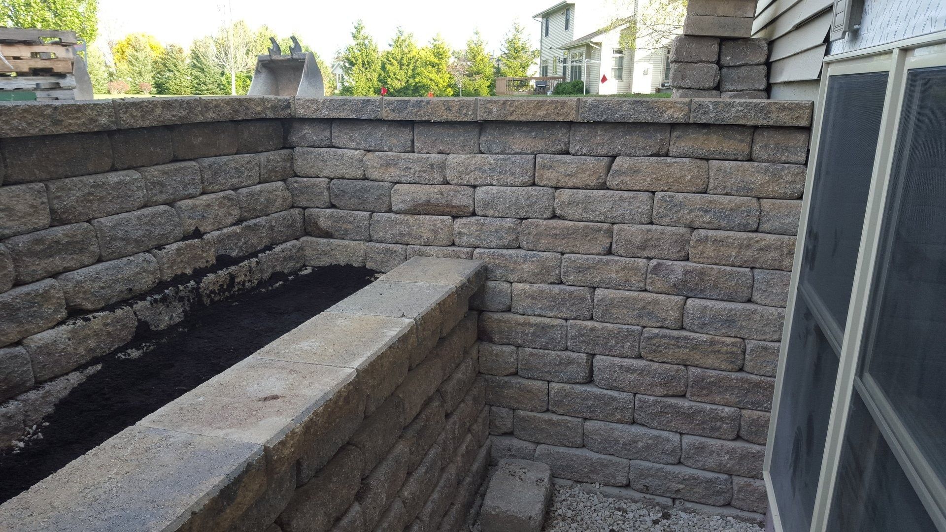 A tiered stone retaining wall planter cornered against the side of a house with dark soil inside.
