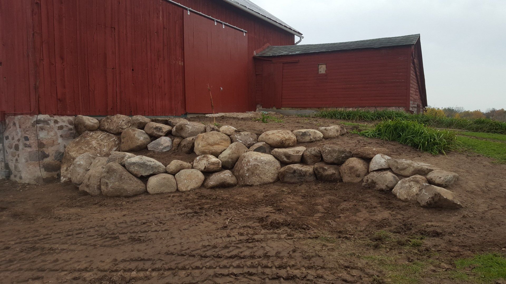 A red barn with a stone retaining wall built into the sloped, muddy ground in front of it.