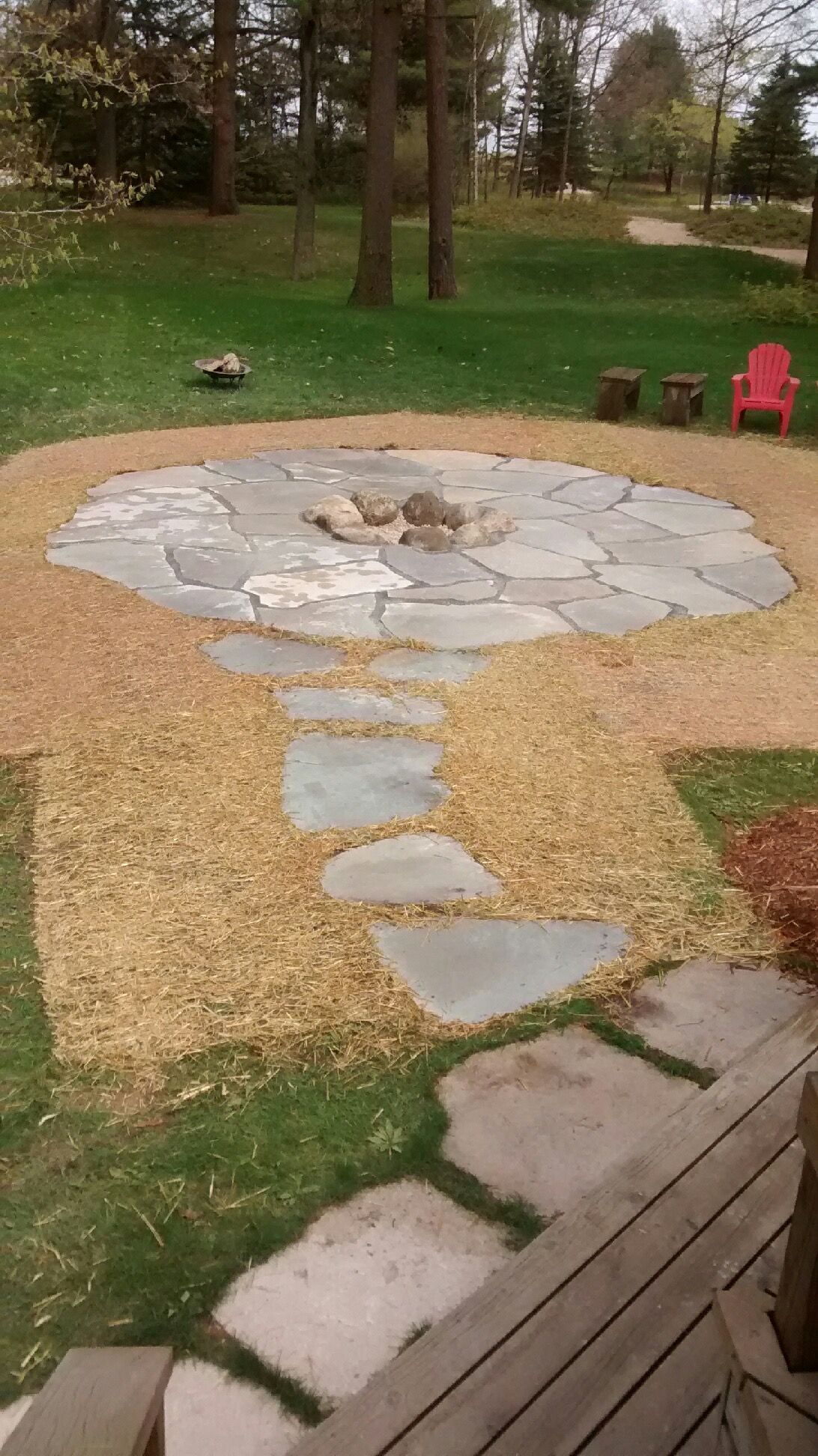 A backyard fire pit area with stone pavers surrounded by wood chips, leading to a wooden deck, set against a lawn.