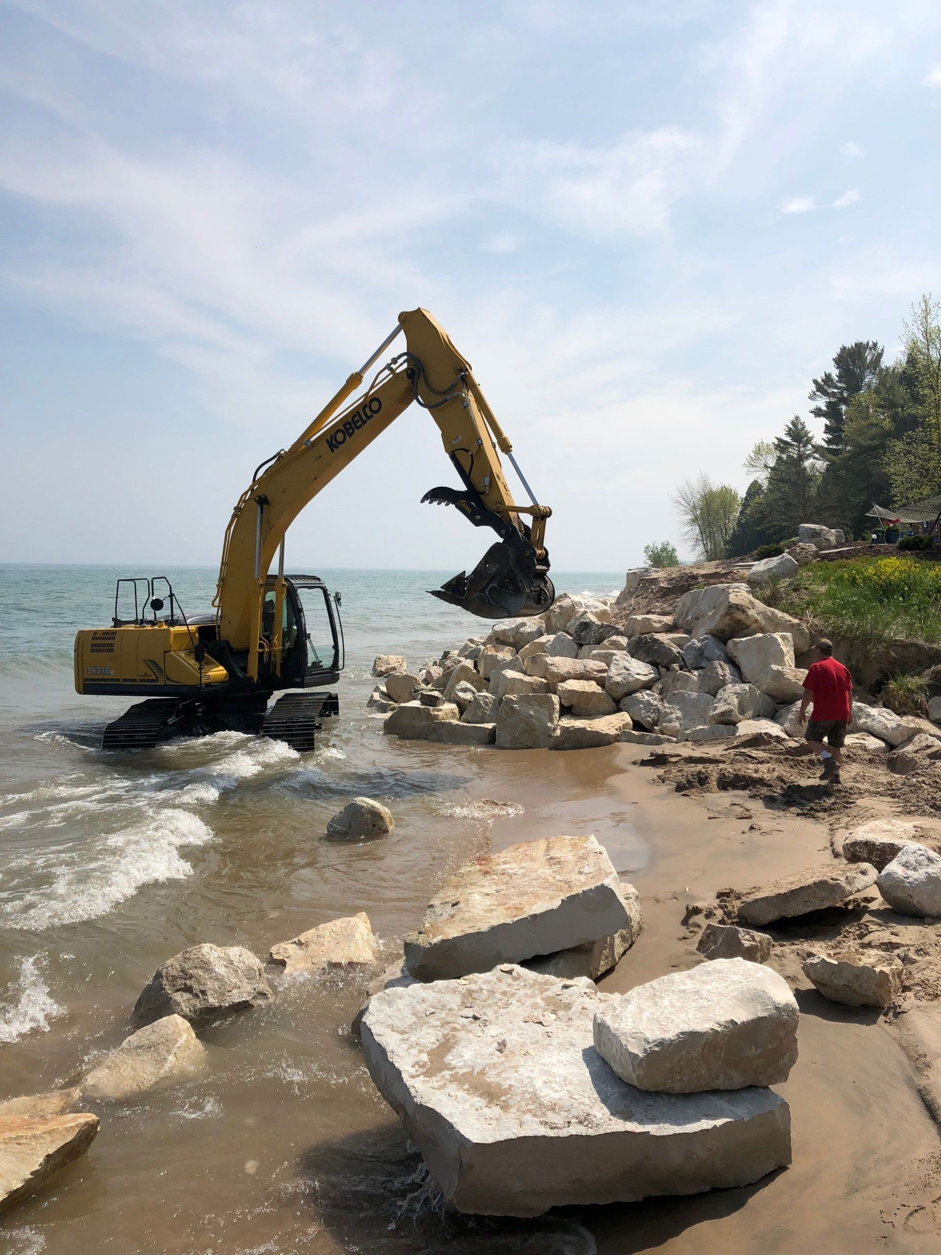 A yellow excavator places large rocks along a lakeshore, as a person stands nearby on the beach.