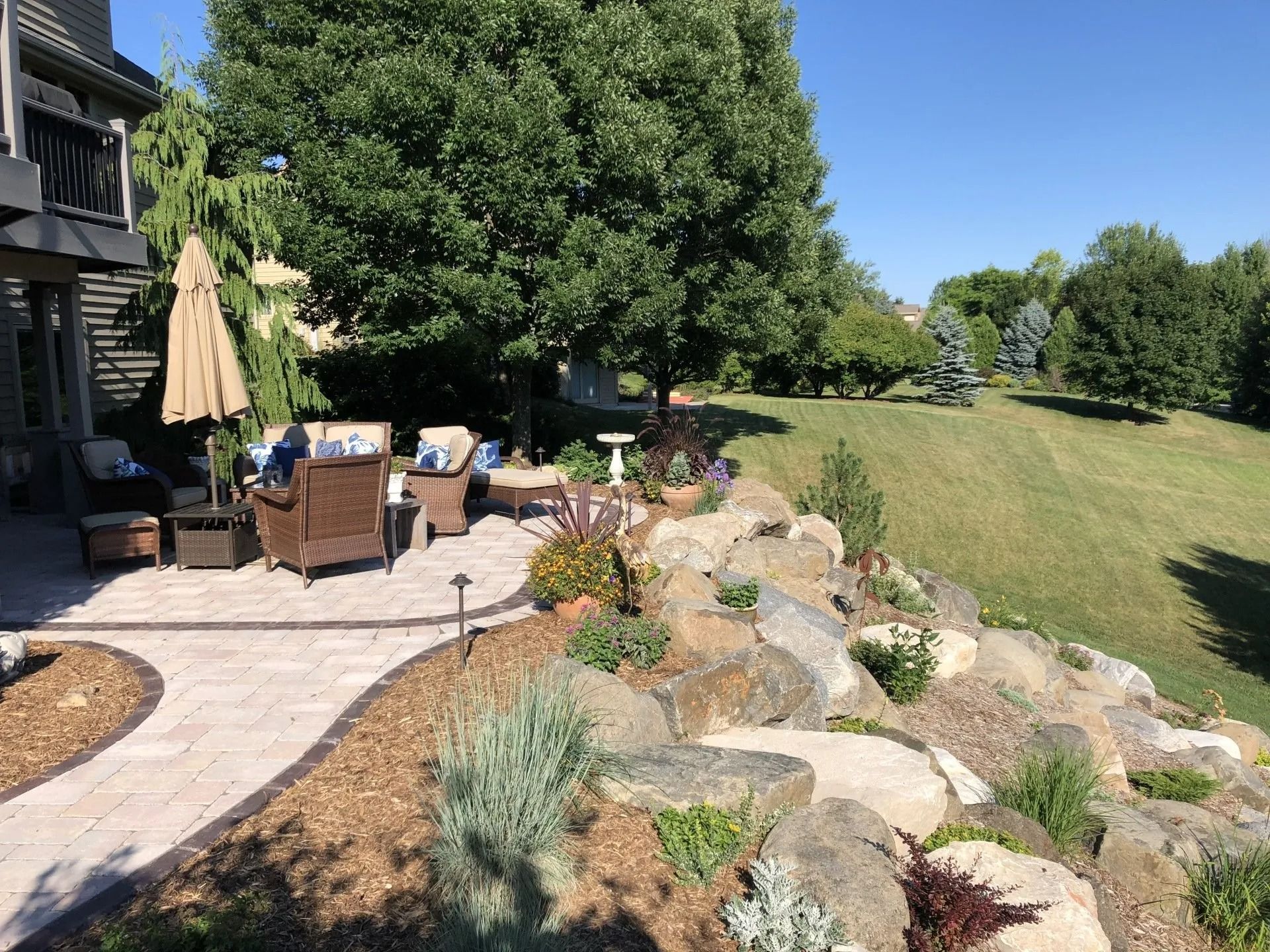 A stone patio with outdoor furniture overlooks a landscaped rock garden and a large, grassy backyard on a sunny day.