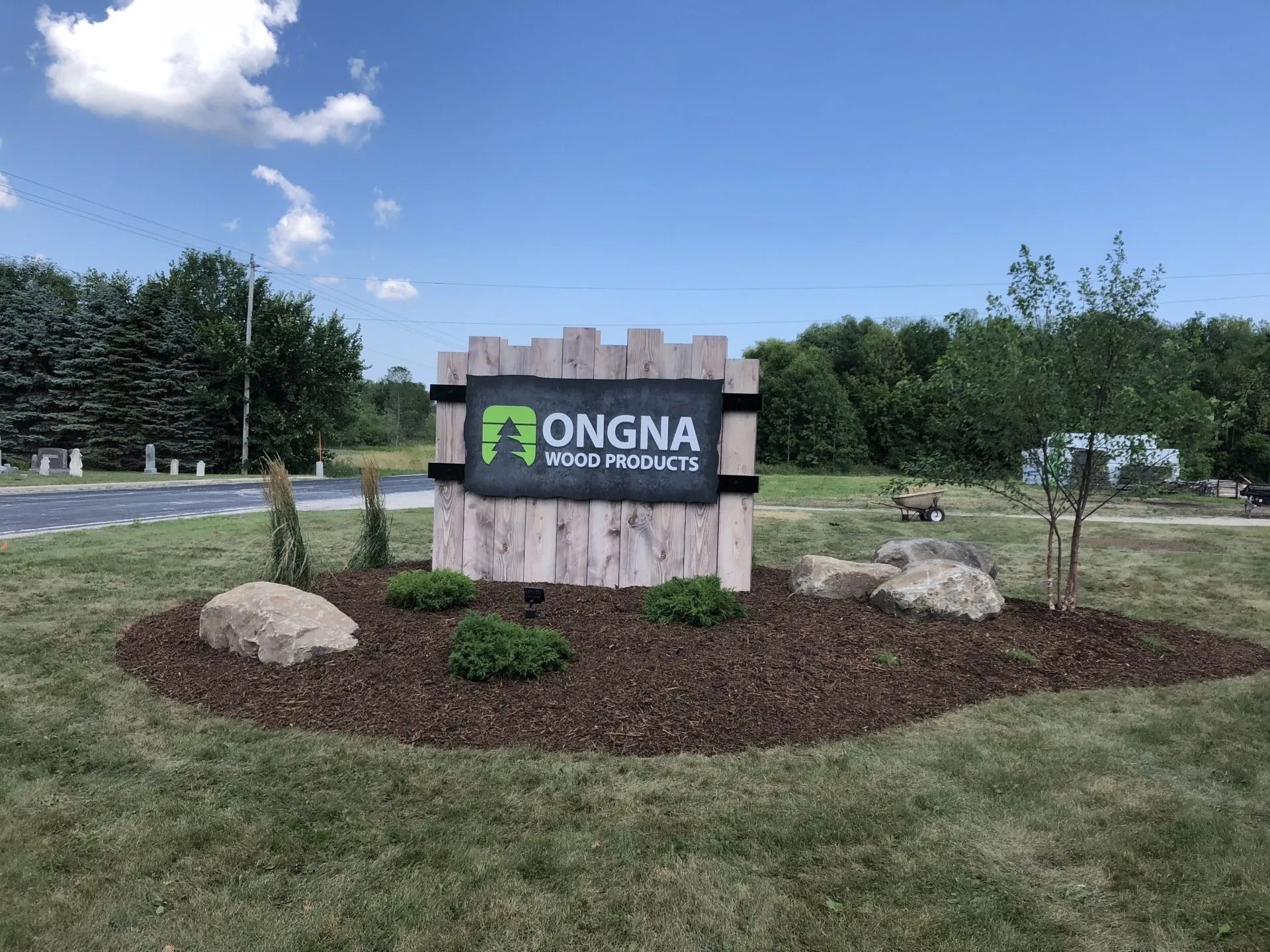 A stone and wood sign for Ongna Wood Products stands in a mulched garden bed under a bright blue sky.