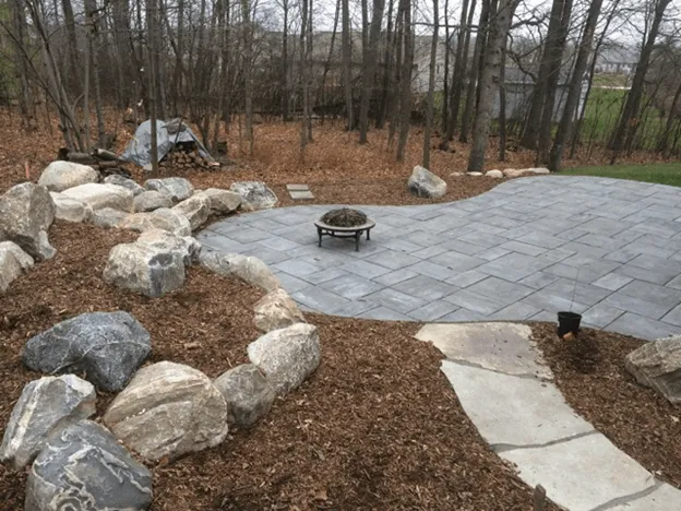A grey paver patio with a fire pit, surrounded by large landscaping rocks and a flagstone path in a wooded backyard.