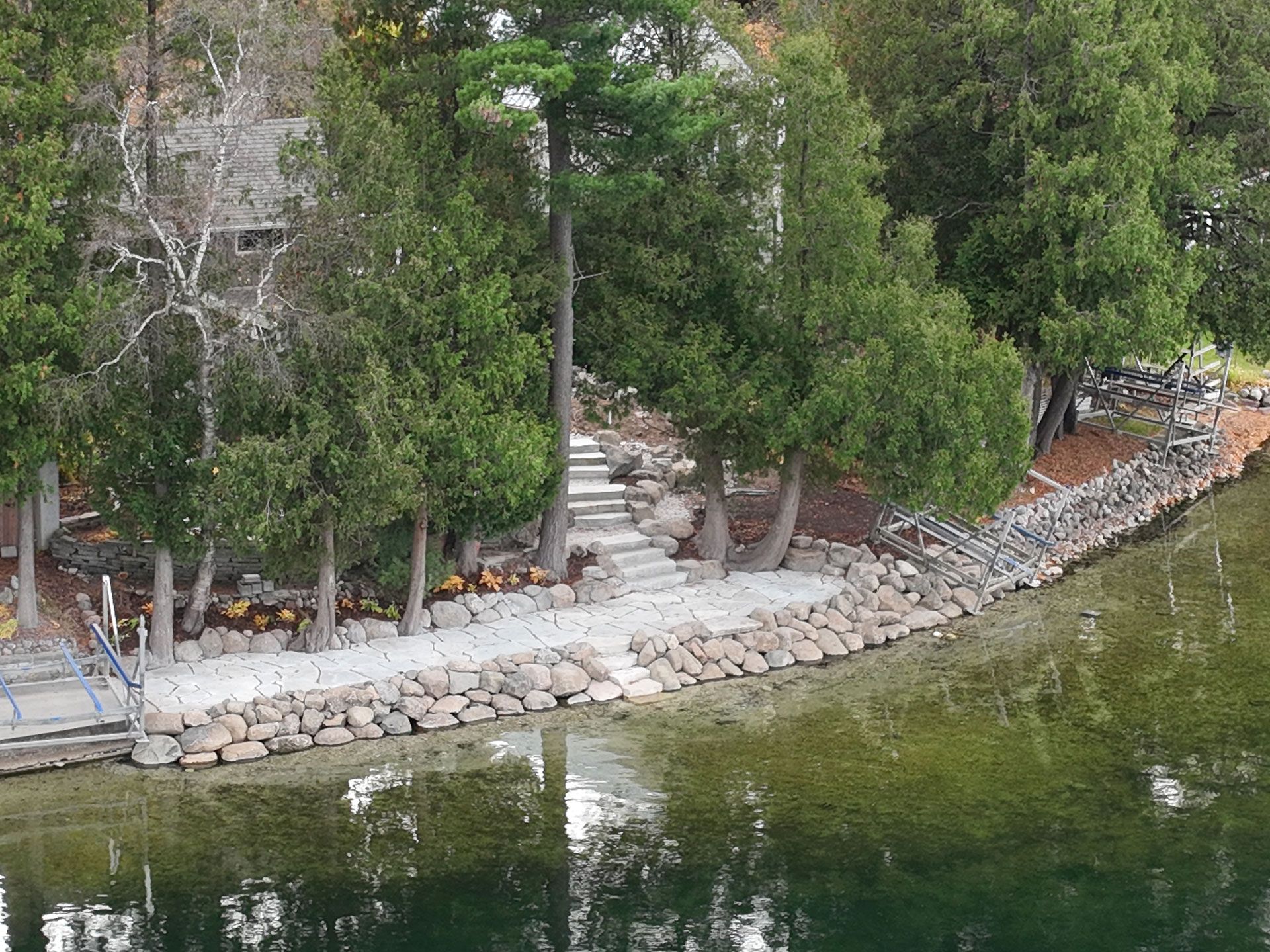 A stone retaining wall lines a lakeshore, leading to stairs set among trees leading toward a house.