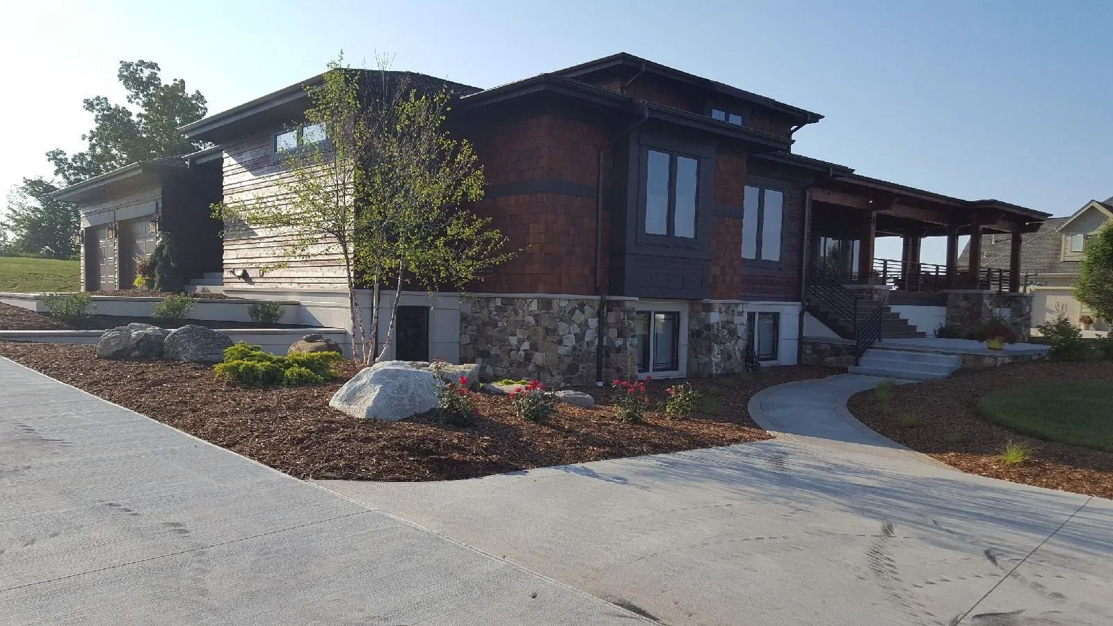A modern home with wood and stone siding, a covered porch, a concrete driveway, and landscaped front yard.