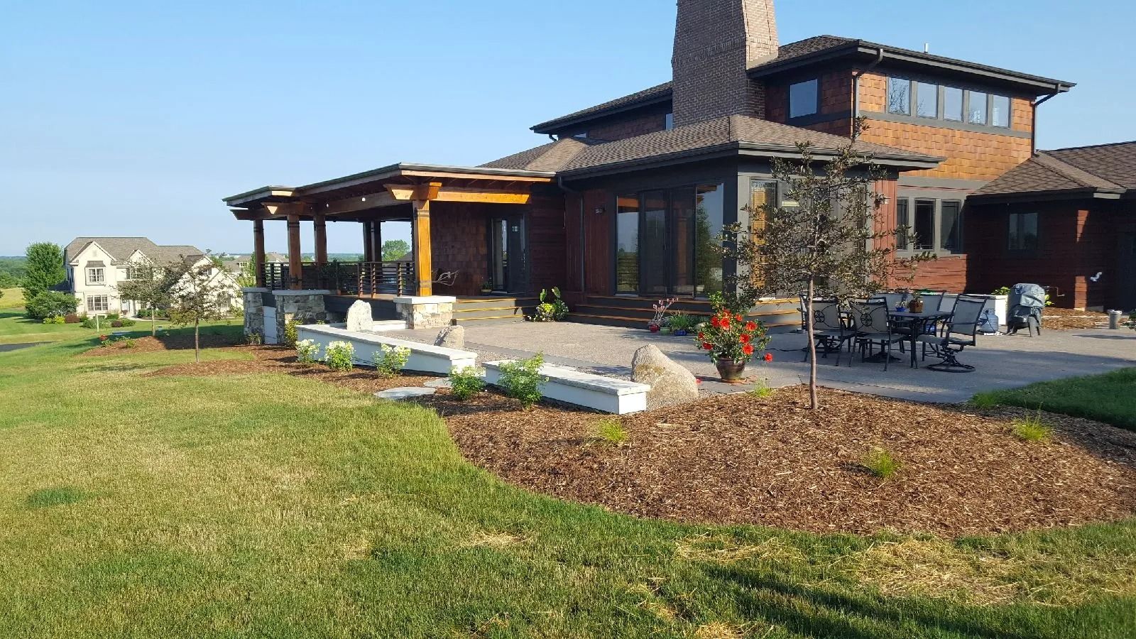A multi-story brown house with a stone chimney, a covered patio, and a landscaped yard with mulch beds and green grass.
