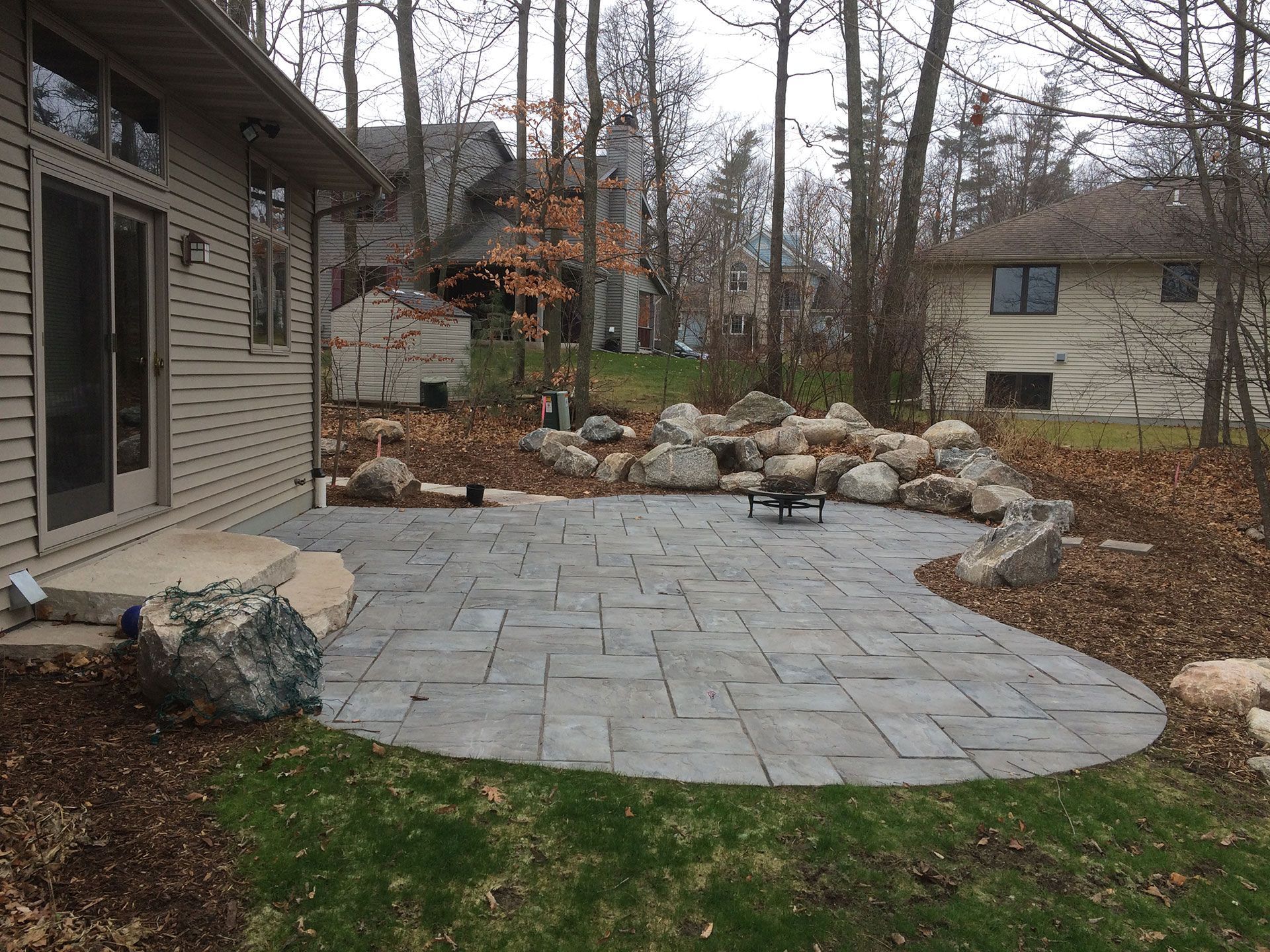 A backyard patio made of grey rectangular pavers, bordered by large decorative boulders and a wooded area.