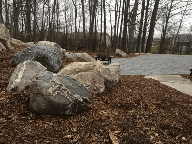 Large, grey boulders sit in a mulch bed next to a paved stone patio, with a wooded area in the background.