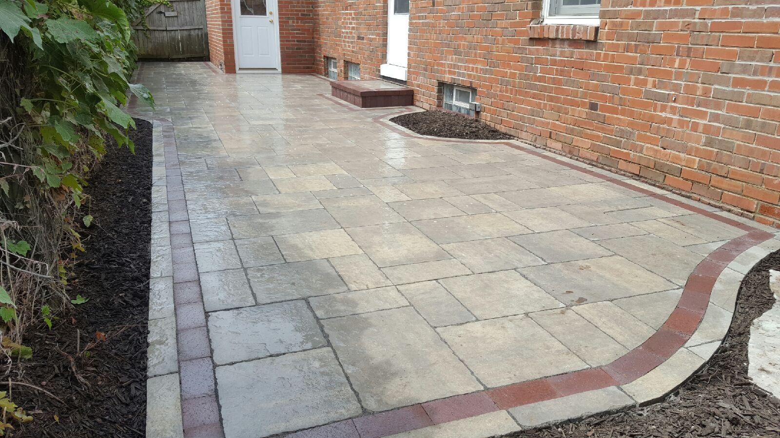 A newly installed patio made of light-colored rectangular pavers with a dark red border, set against a brick house wall.