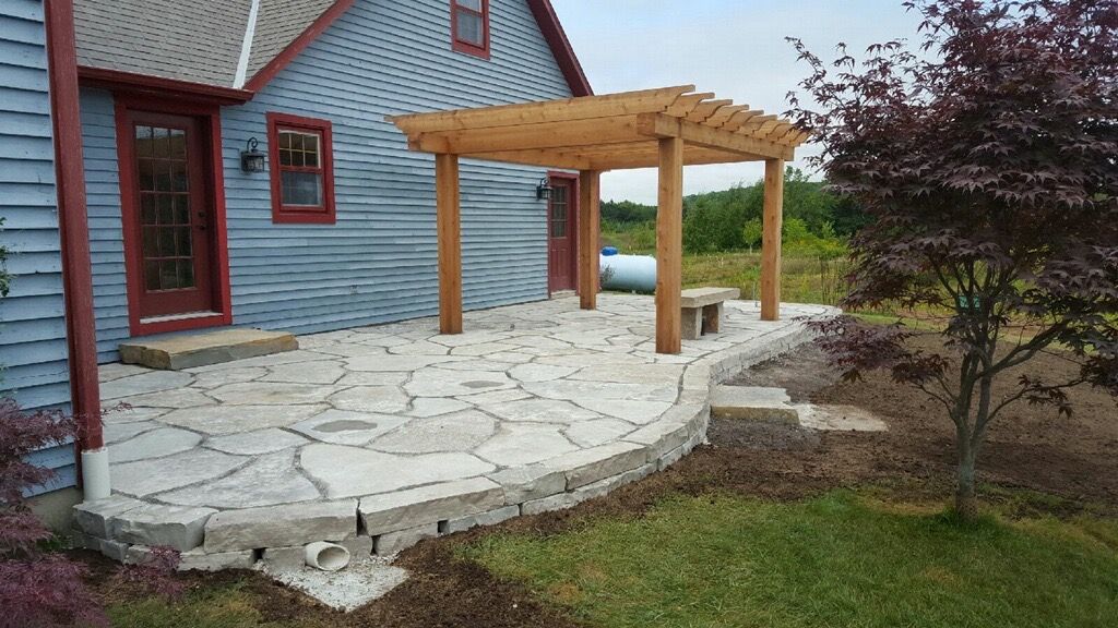 A flagstone patio with a wooden pergola, situated next to a light blue siding house, with a small tree in the yard.