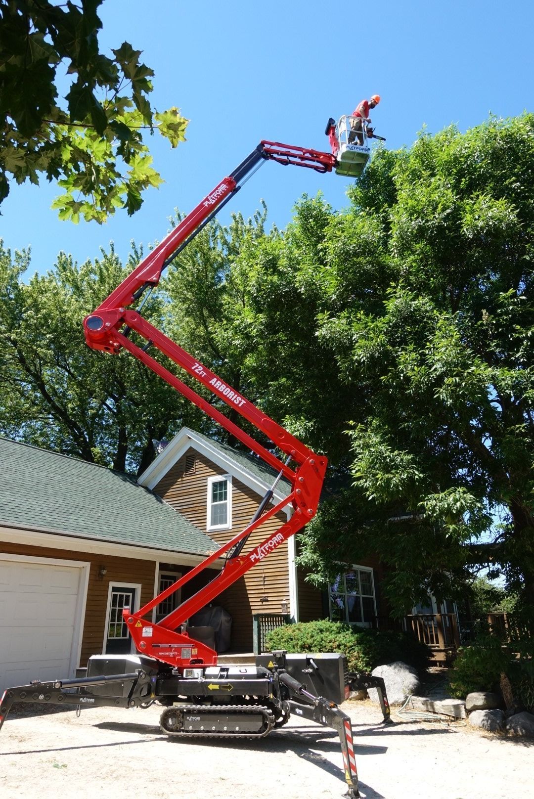 A red compact tracked lift extends its articulated boom high into a tree next to a house on a sunny day.