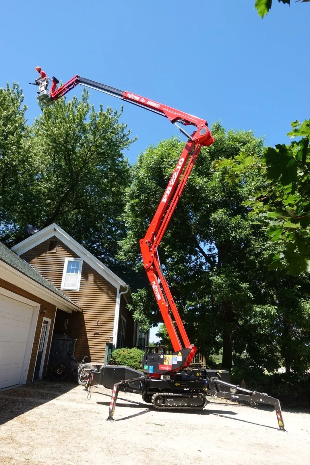 A person in a red cherry picker lift trimming tree branches next to a house on a sunny day.