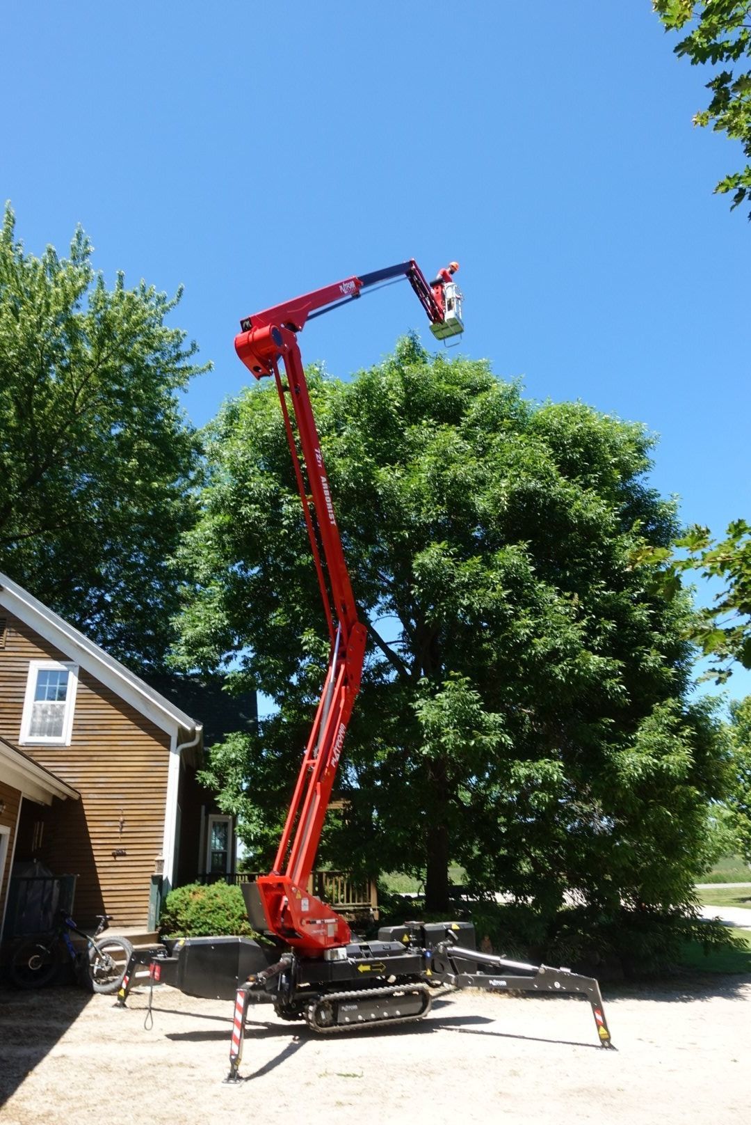 A red track-mounted articulated boom lift stands extended near a tree and a house on a sunny day.