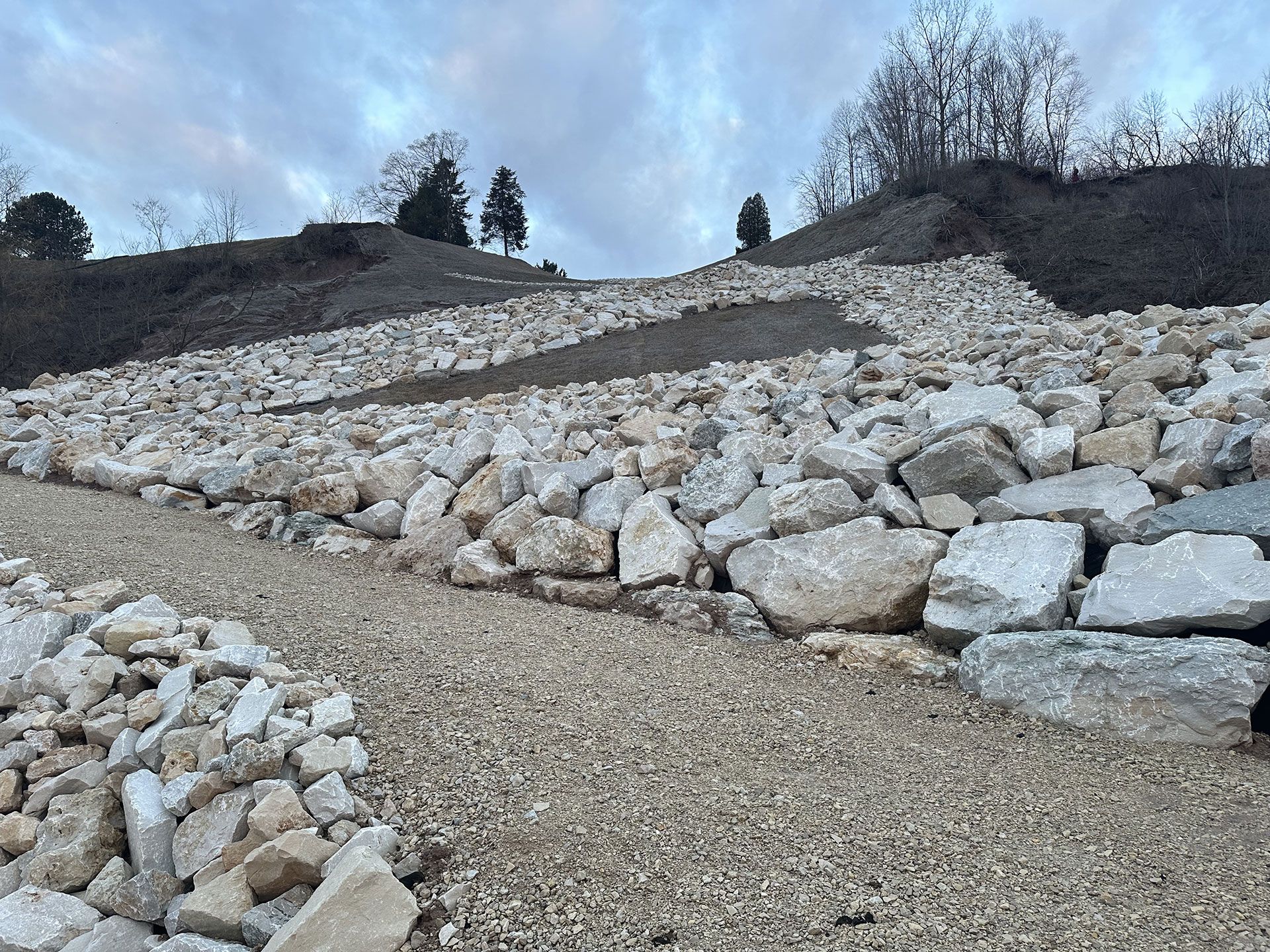 Large boulders line a sloped embankment next to a gravel path against a cloudy sky.