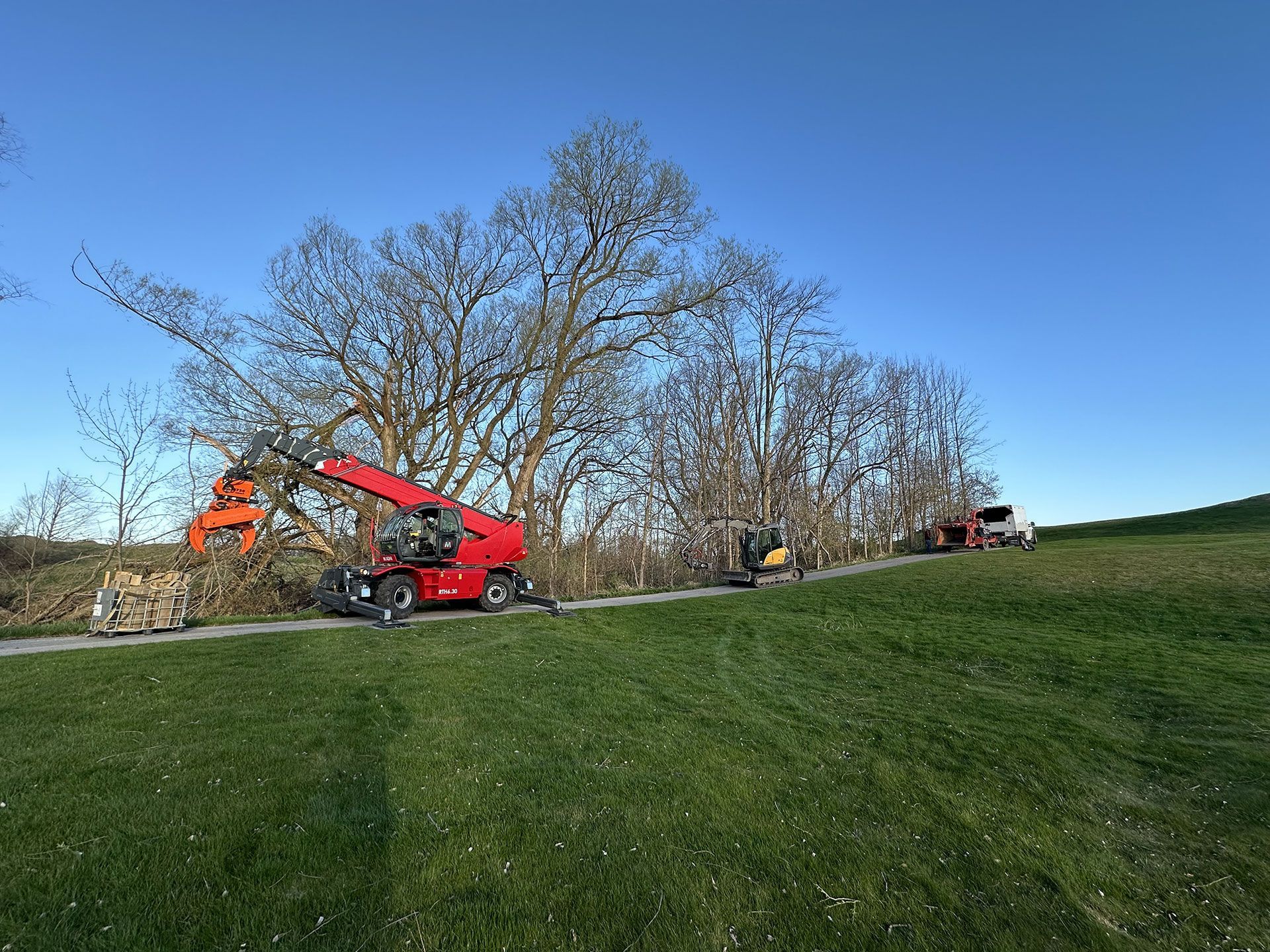 A red telehandler and other construction machinery work on trimming trees along a path in a grassy field.