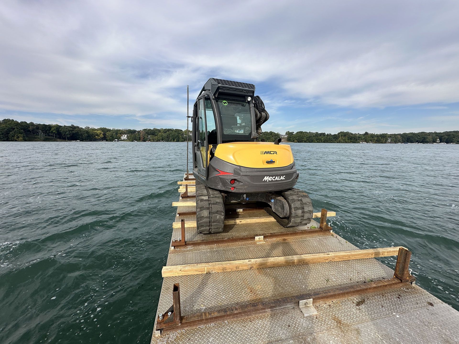 A yellow and gray excavator sits on a metal barge floating on a lake under a cloudy sky.