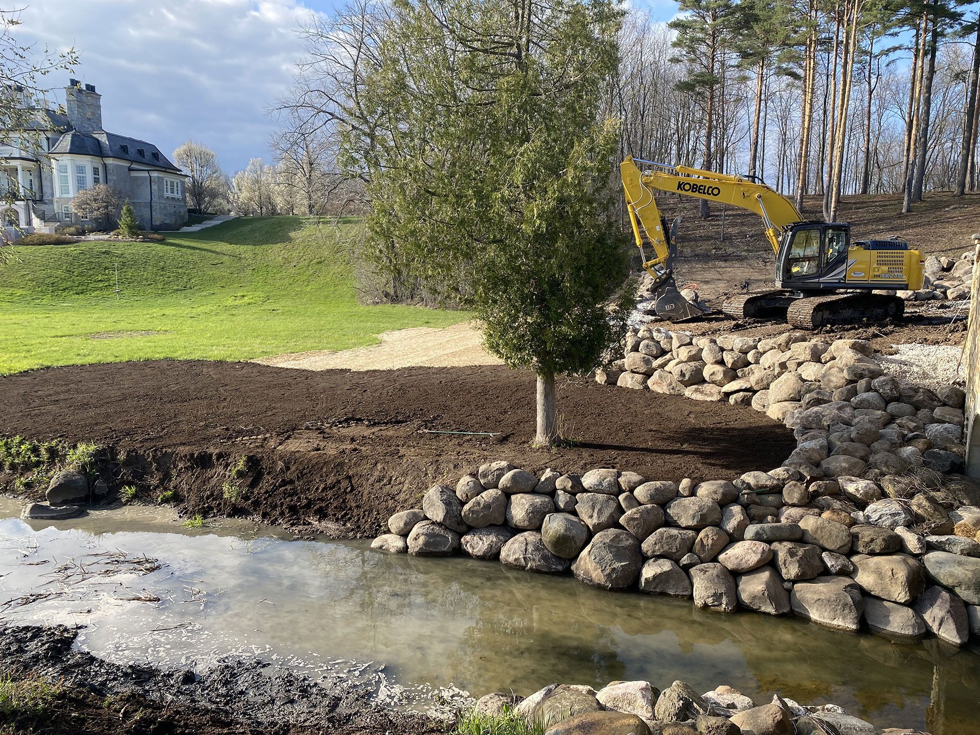 A yellow excavator parked above a rocky bank lining a stream, with a grassy landscape and large estate house nearby.
