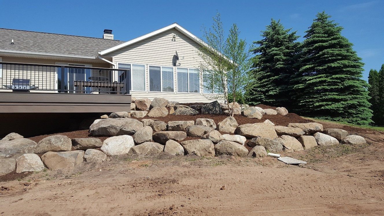 A stone retaining wall in front of a house with a wooden deck, bare earth in the foreground, and pine trees.