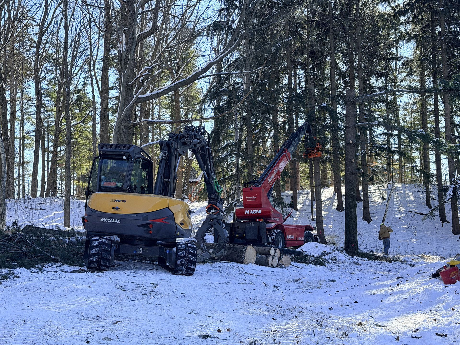 A yellow excavator and a red crane work in a snowy, wooded area to clear fallen logs.
